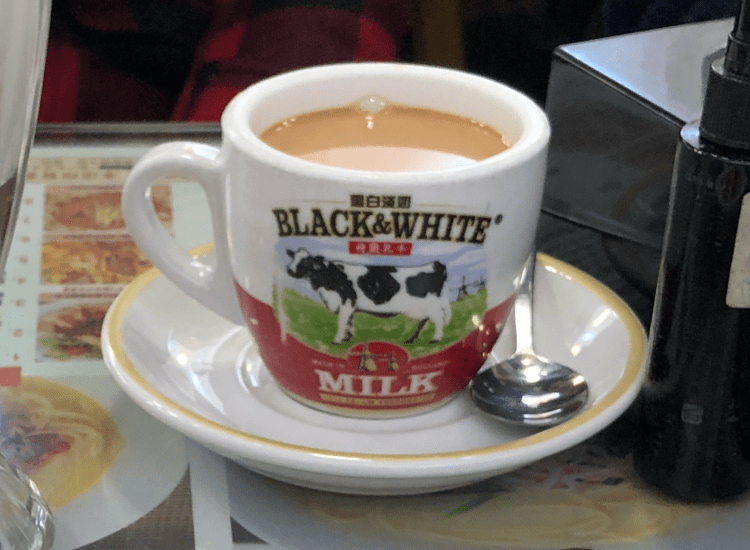 一杯裝有黑白牛奶的咖啡，杯子上印有奶牛圖案，旁邊有一個小湯匙和盤子。